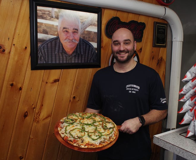 Supreme House of Pizza owner Manny Lambrakis, on Tuesday, Nov. 14, 2023, holding a pizza with cheese, green peppers and other ingredients, stands next to a picture of his dad, Jimmy Lambrakis, who founded the restaurant in 1972 and worked in it right up to his passing in 2016.